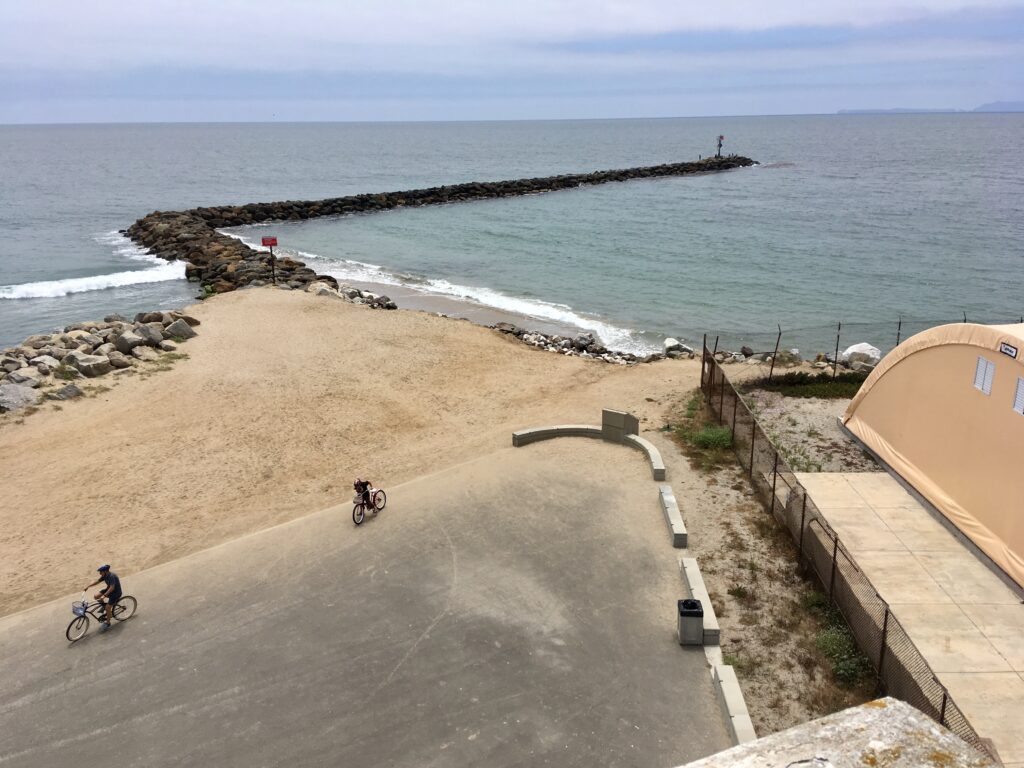 Coastal scene with rocky breakwater, sandy beach, and two cyclists on a paved path near a beige structure, with a lighthouse visible in the distance. Caption: Lighthouse surroundings, 2019. Credit: Craig Baker.