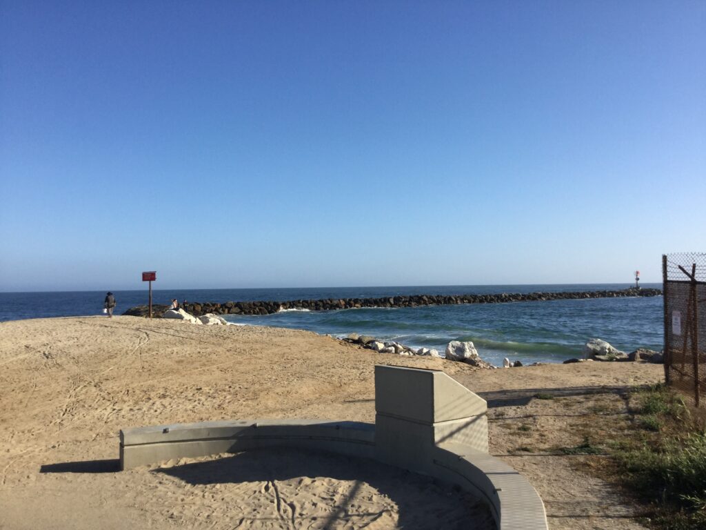 Sandy beach with concrete ramp, red sign, and distant lighthouse. Rocky breakwater, clear blue sky, and person walking near ocean.