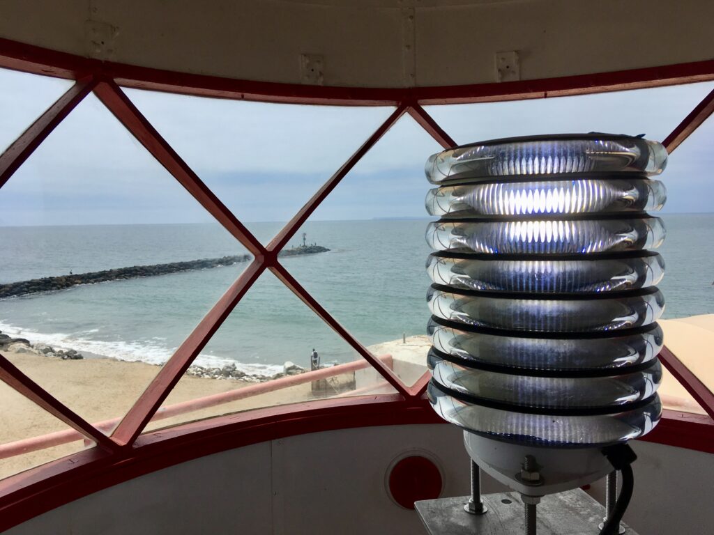 A multi-ring navigational light inside a lighthouse with red metal framing, overlooking a sandy beach, rocky breakwater, and ocean under an overcast sky. Photograph from Lightworks 2019 by Craig Baker.
