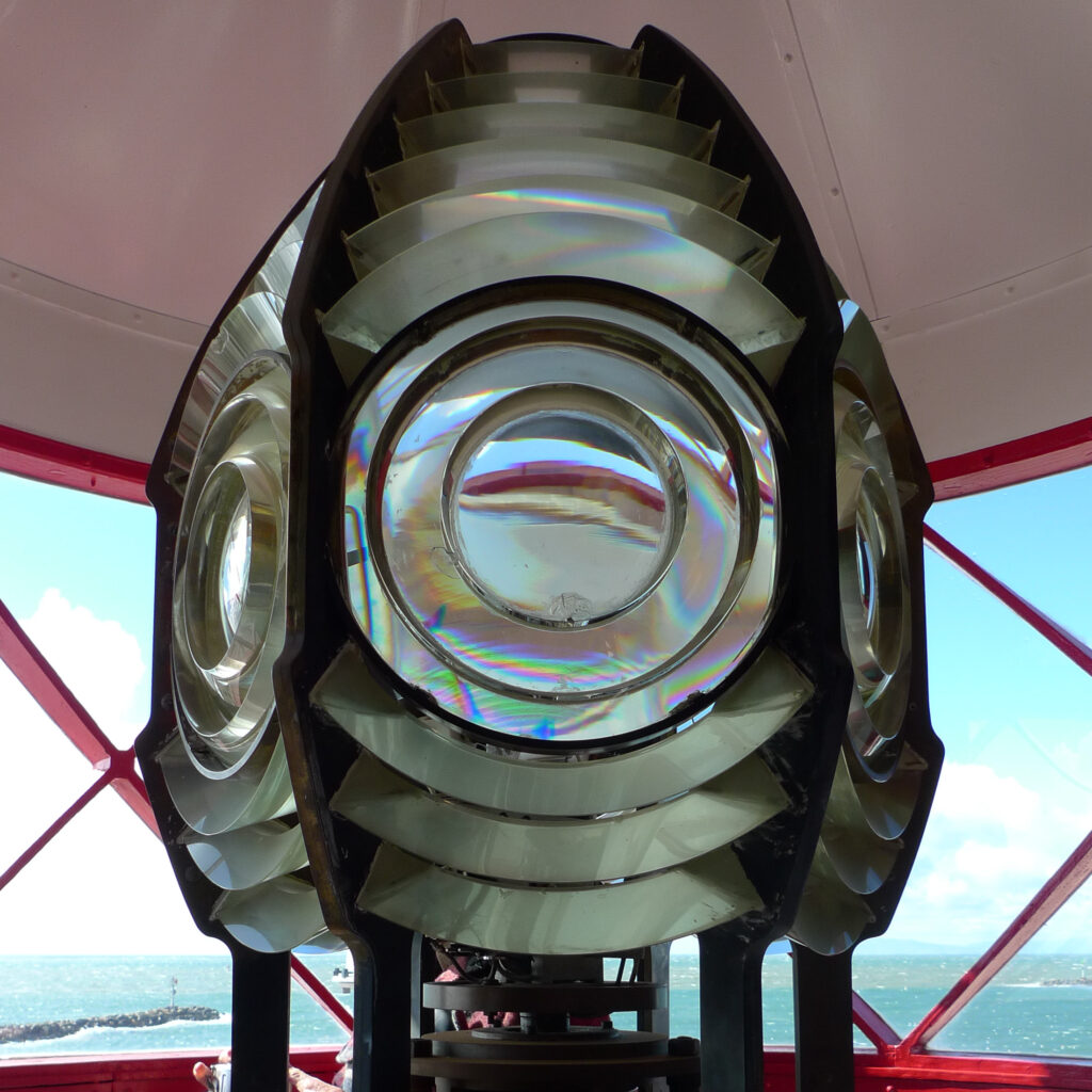 Close-up of a lighthouse Fresnel lens showing iridescent rainbow reflections, with the ocean visible through red-framed windows. Caption: Lightworks by Stephen Schafer.