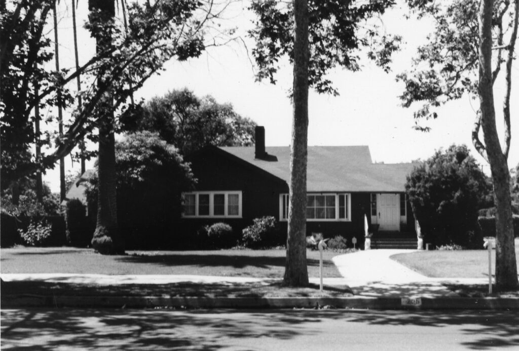Murphy House, a single-story dark house with white-framed windows and a white door, surrounded by trees and a lawn, circa 1980s.