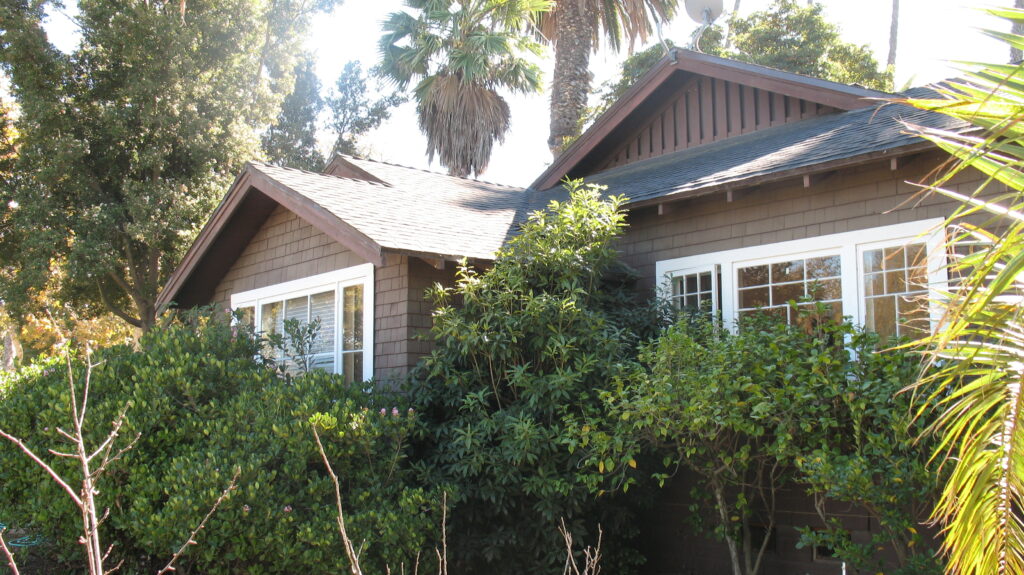 Brown shingle house with white-framed windows, surrounded by dense green shrubs and palm trees, photographed in 2011 as part of Murphy House details.