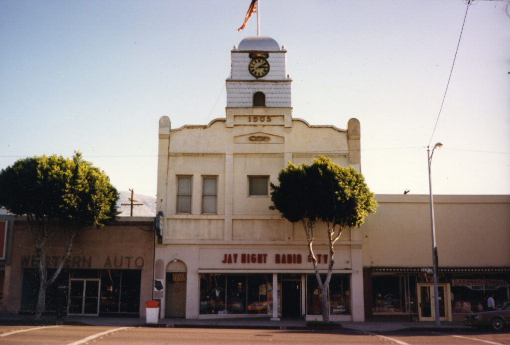 Beige Odd Fellows' Town Clock building with a clock tower marked "1905", featuring "JAY NIGHT RADIO TV" storefront, flanked by "WESTERN AUTO" and "BARGAIN BAZAR" businesses, surrounded by trees and a street.