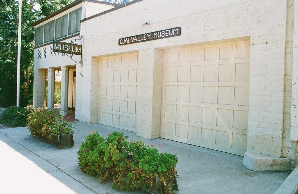 White brick building with "OJAI VALLEY MUSEUM" signage above garage doors, former County Fire Station No. 21 (1985), featuring planters with greenery on the sidewalk.