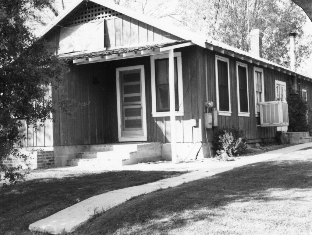 Black-and-white photograph of a 1980s single-story wooden ranch house with a porch, steps, multiple windows, an exterior air conditioner unit, and a sidewalk leading to the entrance.