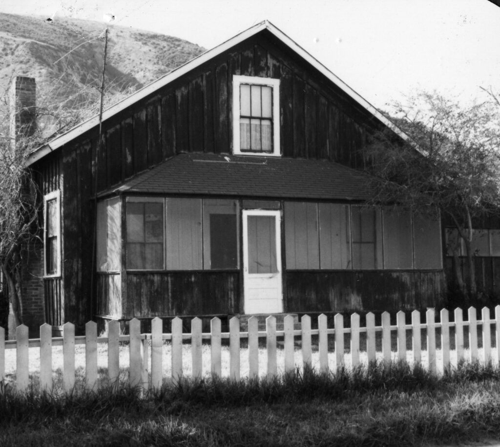 A weathered wooden ranch house with a white picket fence, gabled roof, and mountain backdrop, circa 1980s.
