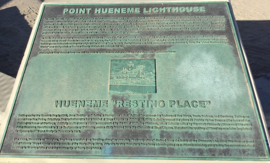 Greenish commemorative plaque with engraved text detailing Point Hueneme Lighthouse history and Hueneme's "Resting Place" origin, featuring a small lighthouse illustration on a patina surface.