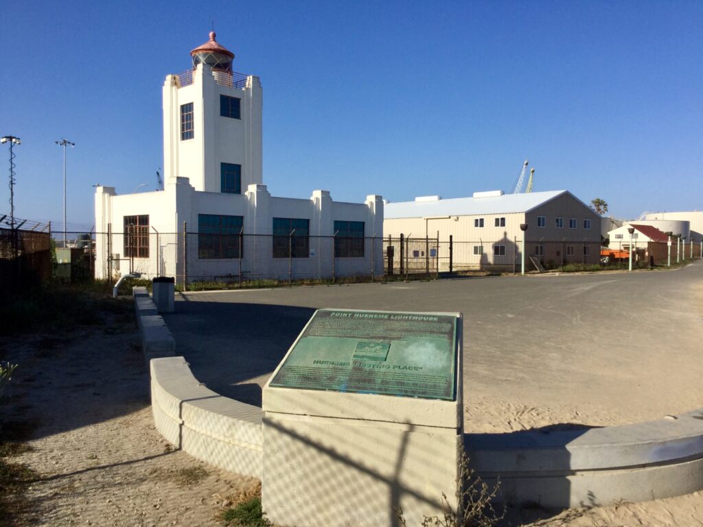 White lighthouse structure with blue windows and red dome tower, surrounded by a fence and parking lot. Foreground features a green plaque reading "POINT HUENEME LIGHTHOUSE" and "HUENEME RESTING PLACE" under a clear blue sky.