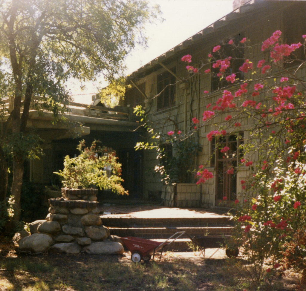 Sunlit garden with pink flowering bushes, stone stack, and red wheelbarrow in front of a two-story Pratt House from the 1980s.