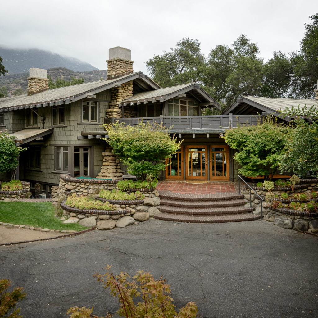 Pratt House exterior with stone chimneys, shingled walls, brick steps to double doors, stone terraces with plants, and mountain backdrop.