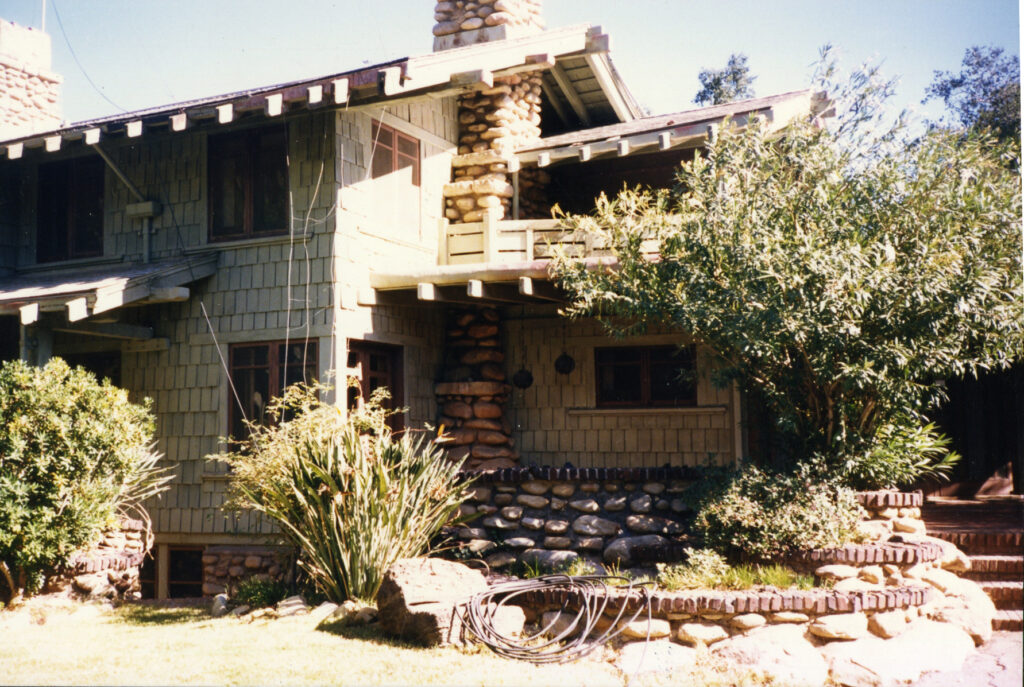 Two-story house with stone and shingle siding, stone chimney, covered porch, and landscaped garden with plants and stone walls. Built circa 1980s.