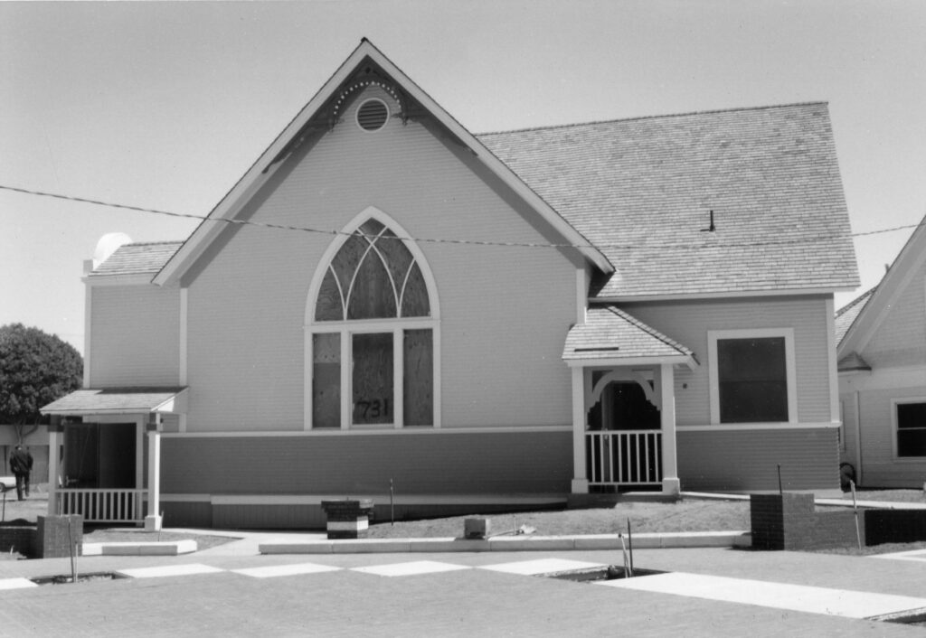 A relocated church building at Heritage Square, circa 1990, features a gabled roof, large arched window with boarded panels displaying '731', and a porch.