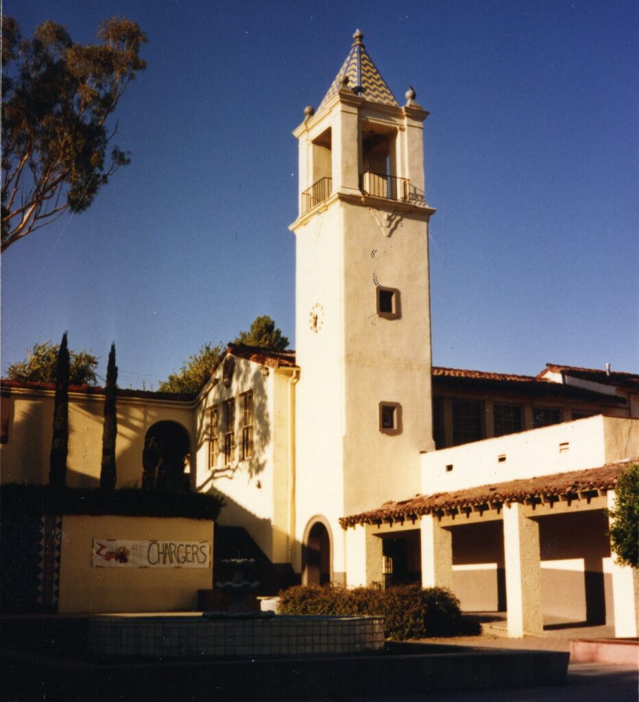 Santa Paula Union High School building from 1985, featuring a beige structure with a clock tower (blue/white tile roof), red-tiled roof sections, and a "CHARGERS" banner near a fountain under a clear blue sky.
