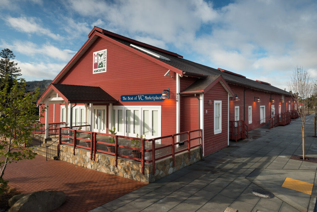 Red building with white trim and stone foundation housing the Museum of Ventura County Agriculture Museum, featuring a blue awning with "The Best of VC Marketplace" sign.