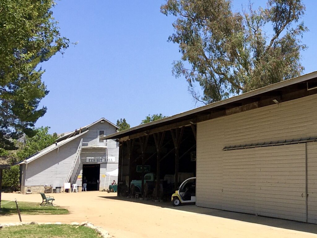 Gray "EXHIBIT HALL" building with a staircase, adjacent to a beige barn with a large garage door; a yellow vehicle is parked under a covered area, surrounded by trees and a dirt path.