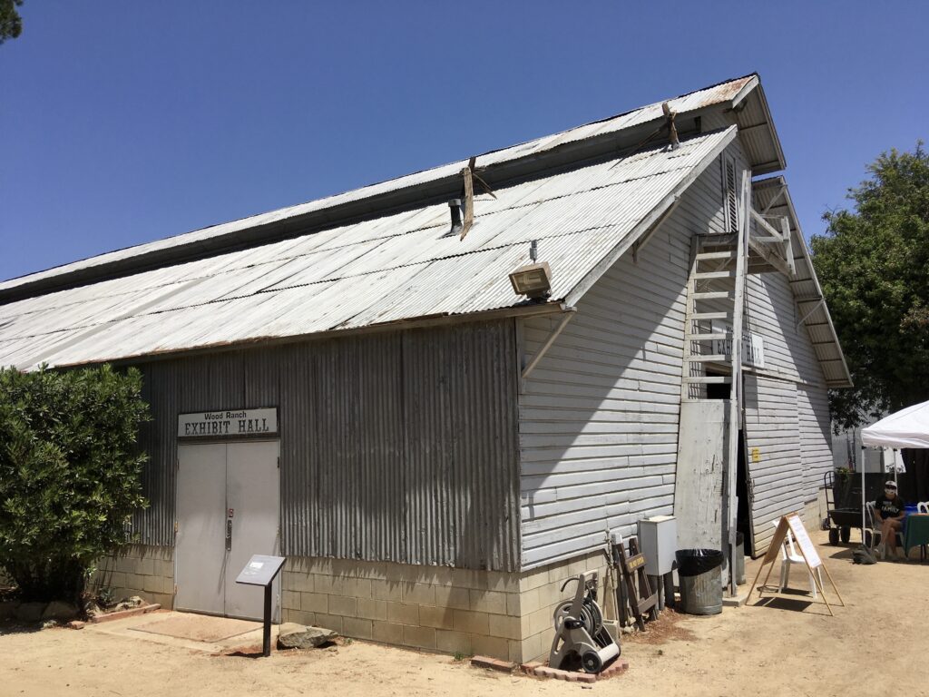 Wood Ranch Exhibit Hall building with corrugated metal roof, white siding, and a ladder on the side. Visible text includes 'Wood Ranch EXHIBIT HALL' on the door, with outdoor elements like a trash can and signboard.