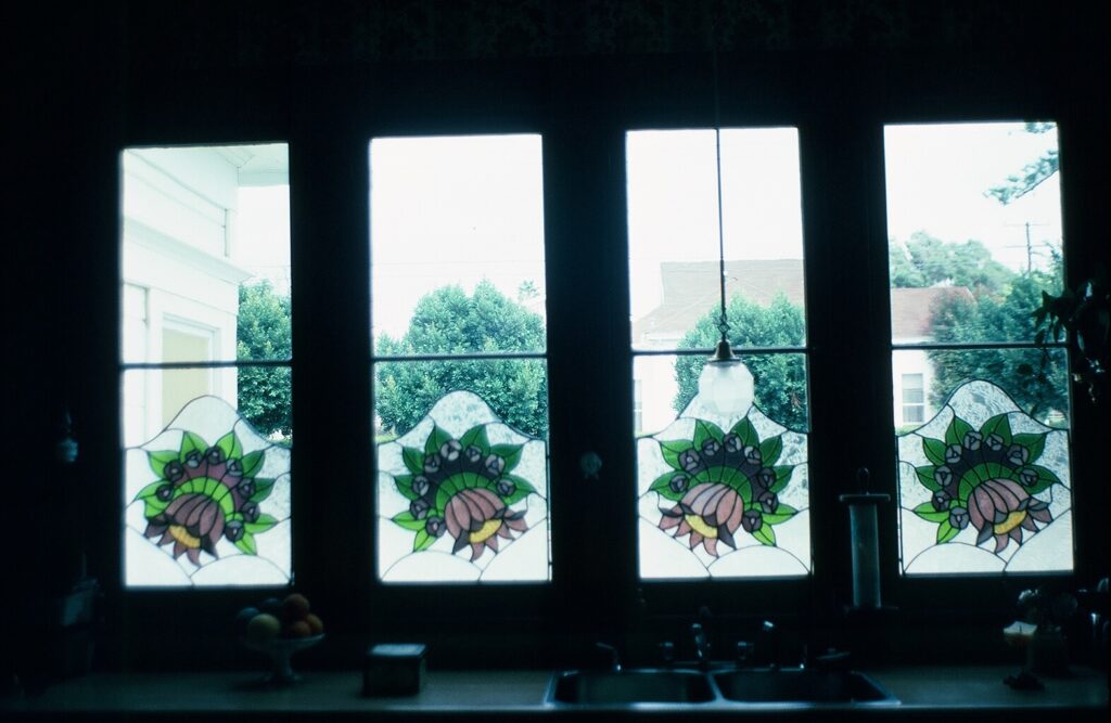 Four stained glass window panes in a kitchen, each with a floral design featuring green leaves, purple centers, and yellow accents, dated 1982.
