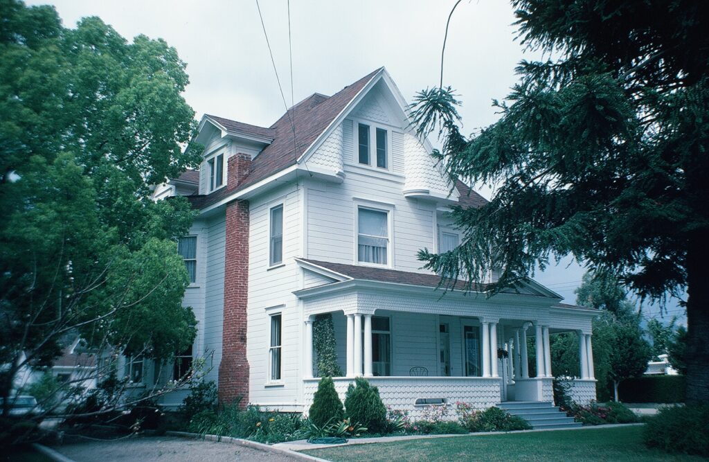 1982 photo of a white Victorian house with a red brick chimney, white porch columns, and green trees surrounding the property.