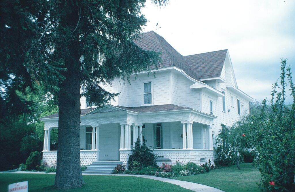 1982 photograph of a white two-story house with a covered porch and brown shingled roof, surrounded by trees and garden. Visible text includes "LARRINGER" on a sign near the left tree.