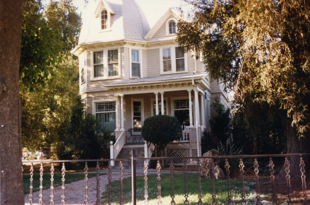 Two-story Victorian house with a porch, columns, and dormer windows, surrounded by trees and a decorative iron fence, circa 1980s.