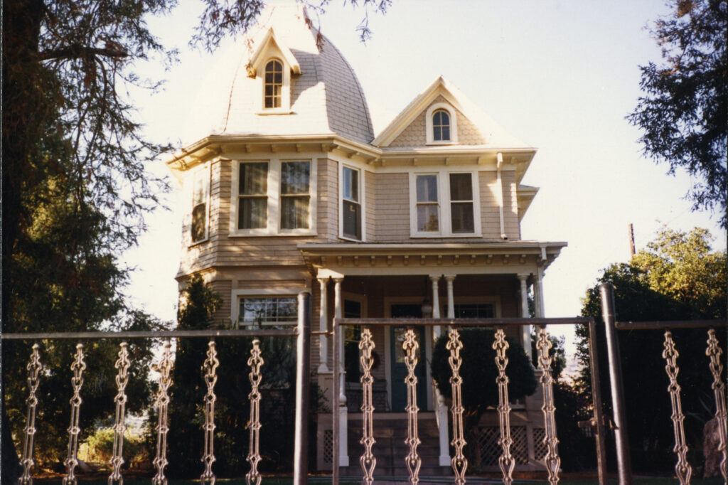 Two-story Victorian house with dome roof, arched windows, and porch, surrounded by trees and ornate metal fence. Light beige siding with white trim and green front door, circa 1980s.