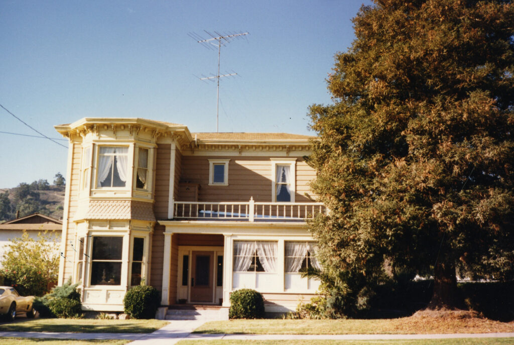 Two-story Victorian house with beige siding, white trim, turret, balcony, and antenna, set against a clear blue sky with a large tree to the right.
