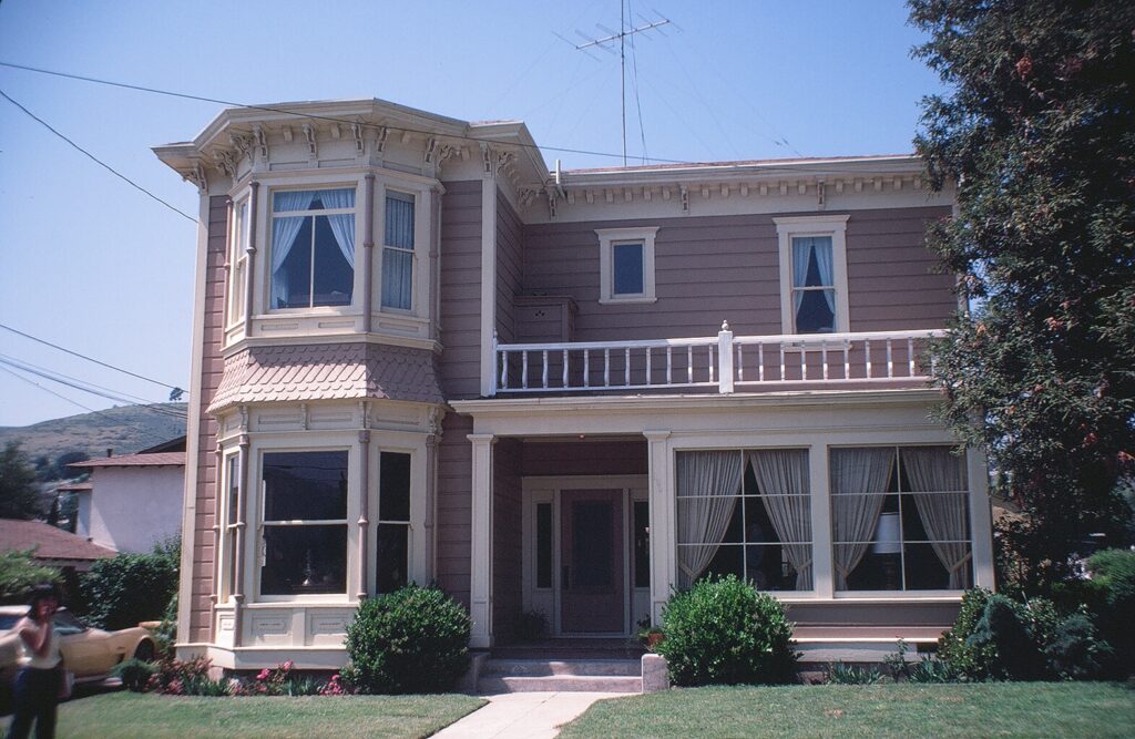 Two-story Victorian house with bay window, balcony, and porch, photographed in 1982. Light brown siding with white trim, surrounded by green lawn and trees under a clear blue sky.