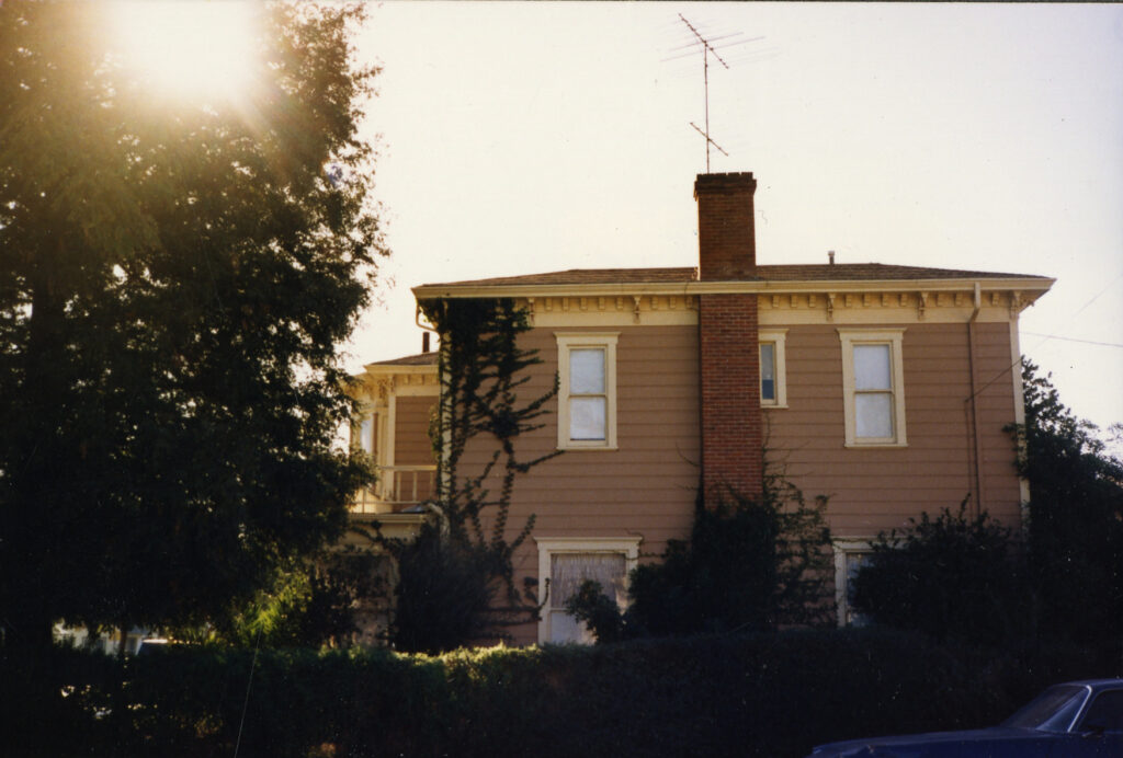 Balcom House, a two-story Victorian home with beige siding, brick chimney, and white window trim, surrounded by trees and a car, circa 1980s.
