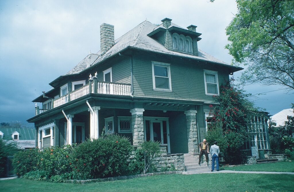 A two-story Victorian-style house with green siding, stone foundation, and white porch columns. Two people stand on the front steps of the Charles Collins Teague House, dated 1982, surrounded by landscaped gardens with bushes and trees.