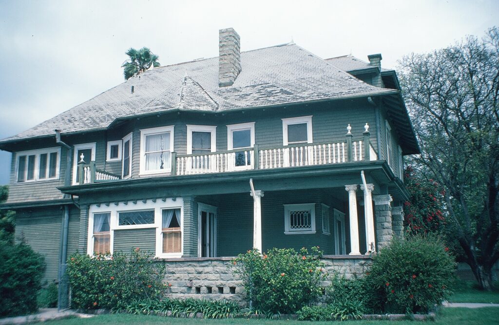 Two-story Victorian house with green siding, white trim, stone foundation, wraparound porch, and second-floor balcony, photographed in 1985.