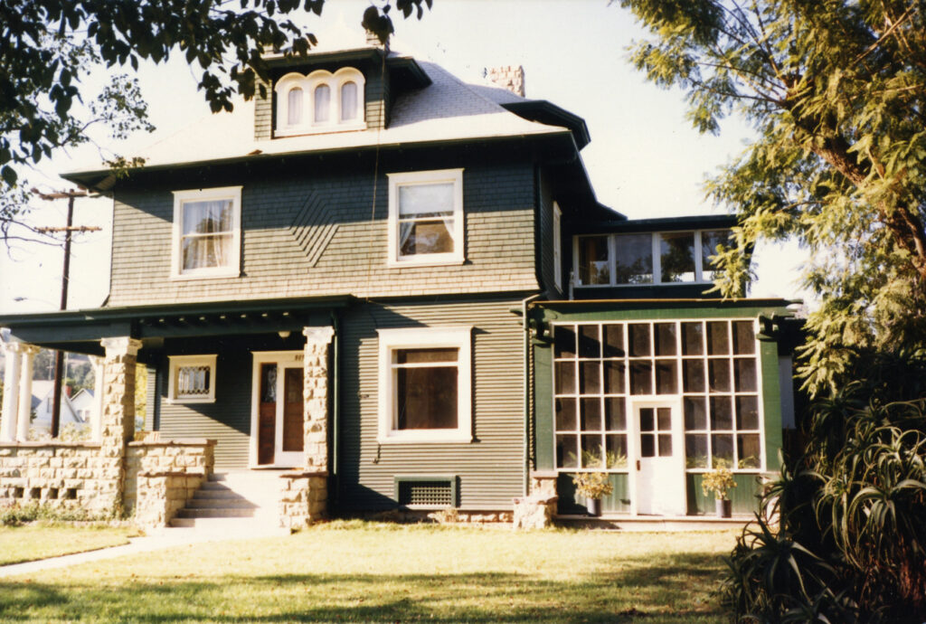Two-story dark green house with white trim, stone columns, dormer windows, and a glass-paned sunroom, circa 1980s.