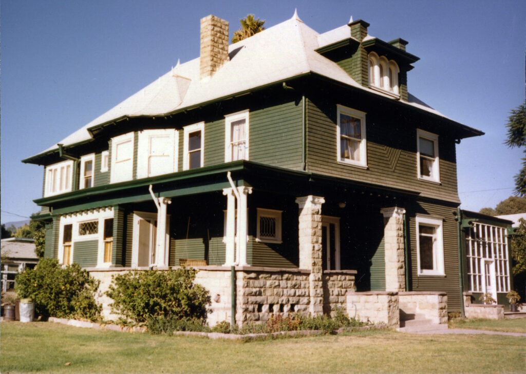 Two-story Victorian house with green siding, white window trim, stone foundation, and wraparound porch, circa 1980s.