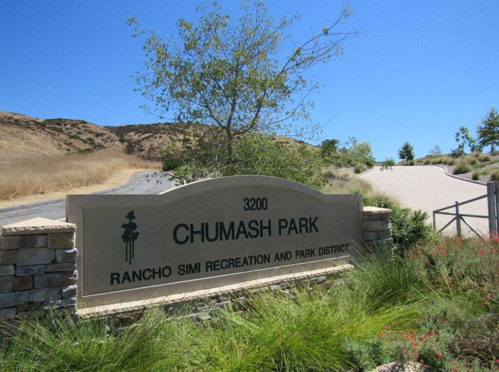 Beige stone entrance sign for Chumash Park (3200) displays "RANCHO SIMI RECREATION AND PARK DISTRICT" text and a tree icon, flanked by stone pillars with a road, hills, and blue sky in the background.