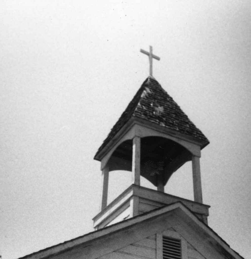 Historical black-and-white photograph of a church steeple with a cross atop a shingled roof and open belfry structure supported by wooden columns.
