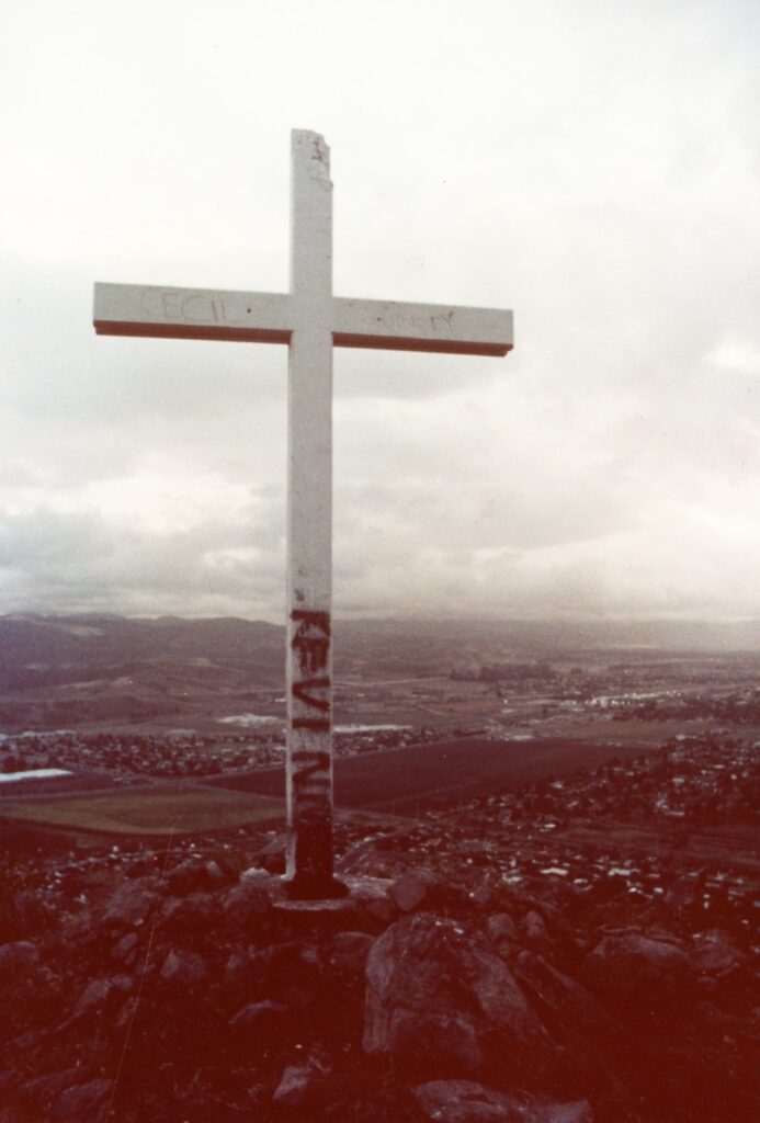 A weathered white cross with graffiti stands atop rocky terrain, overlooking a valley with towns and hills in Simi Valley, circa 1980s.