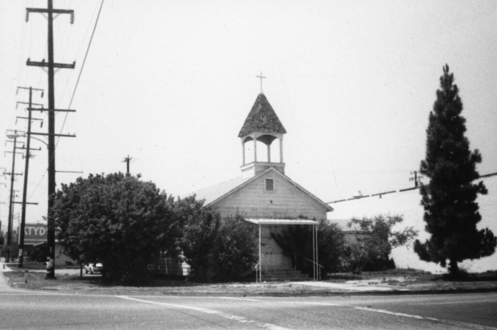 Historical black-and-white photo of a disused church with a steeple and cross, surrounded by trees, power lines, and a street with a partially visible "TYD" sign.