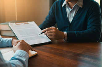 Two people collaborate at a wooden table; one holds a pen over a document clipboard while the other interacts with a notebook.