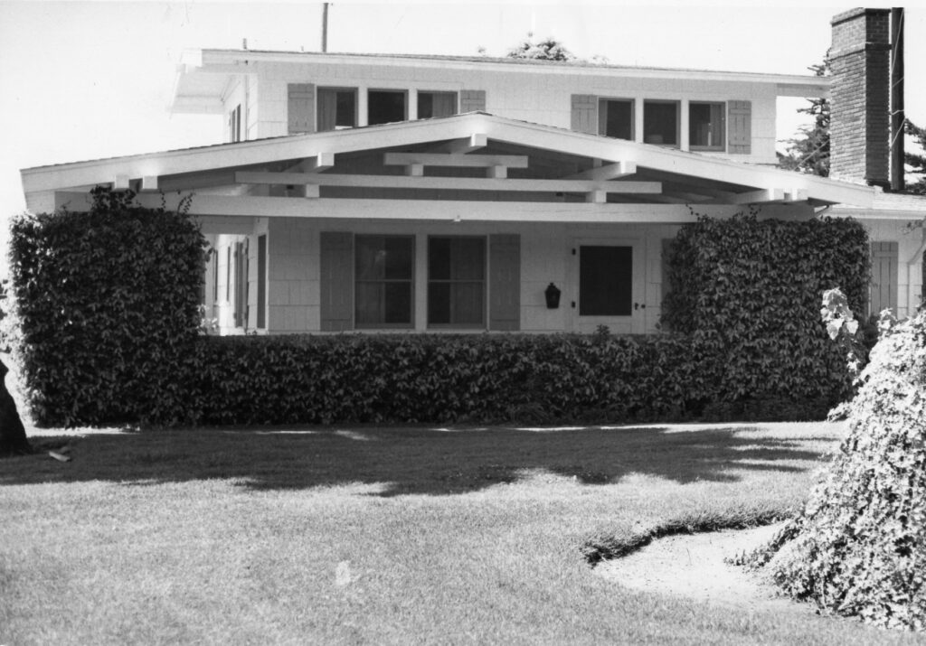 Black-and-white photograph of the Farrell House, a two-story modernist home with a covered porch, hedges flanking the front entrance, and a well-maintained lawn, dated 1996.