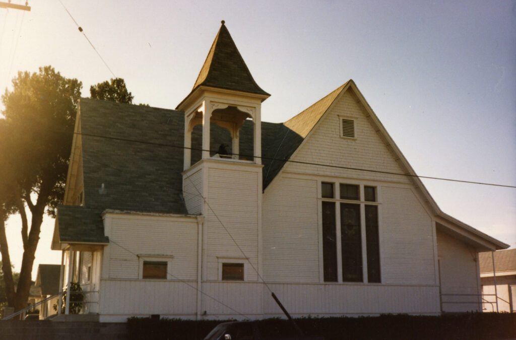 White wooden First Christian Church from the 1980s featuring a steeple, bell tower, and large stained glass window, surrounded by trees.