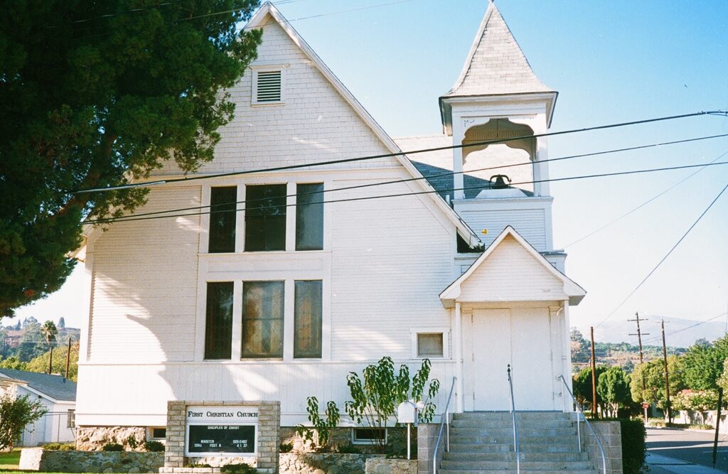 White First Christian Church building with a steeple, bell tower, and entrance steps; sign reads "First Christian Church" with 1985 context.
