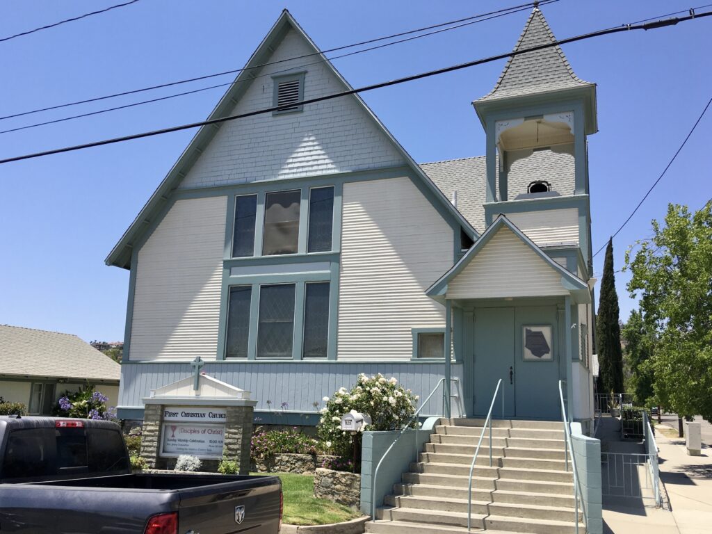 First Christian Church building with light blue and white siding, a bell tower, steps with railings, and a sign reading "First Christian Church (Disciples of Christ)" surrounded by flowers.