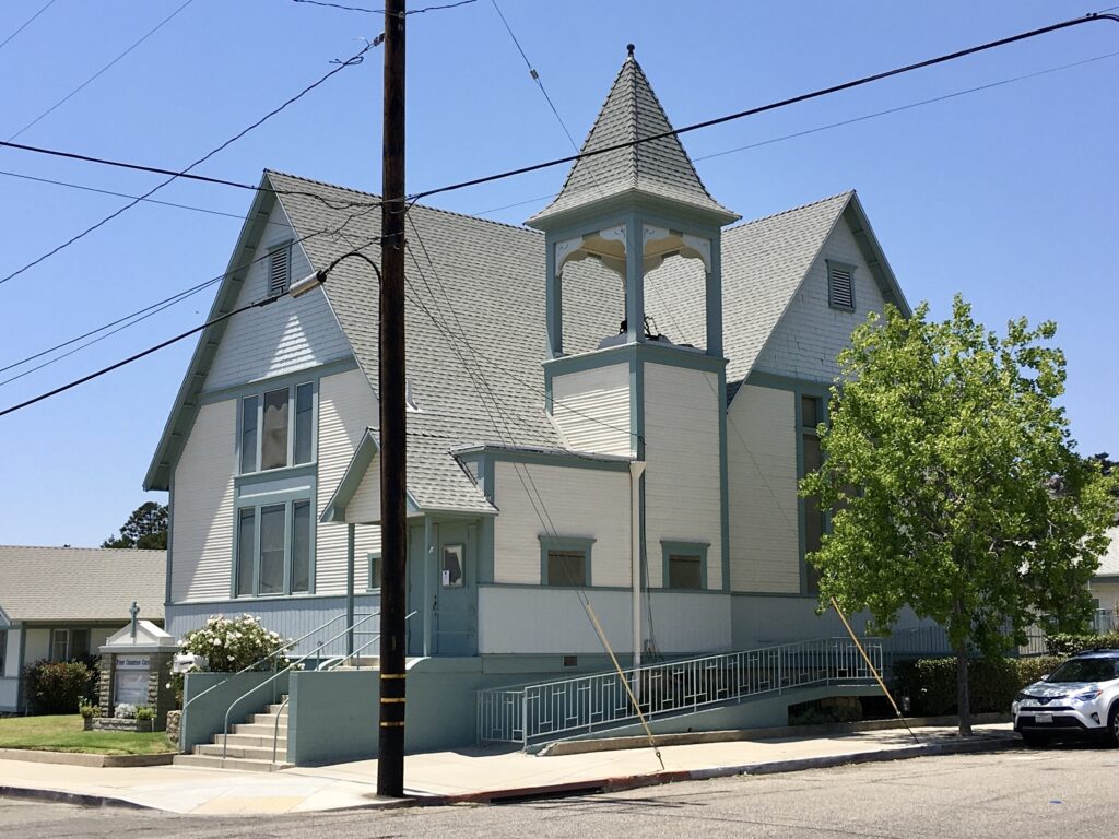 Light blue and white First Christian Church with a central tower, wheelchair ramp, and steps. Features gabled roofs, a utility pole in the foreground, and a white SUV parked on the street under a clear blue sky.
