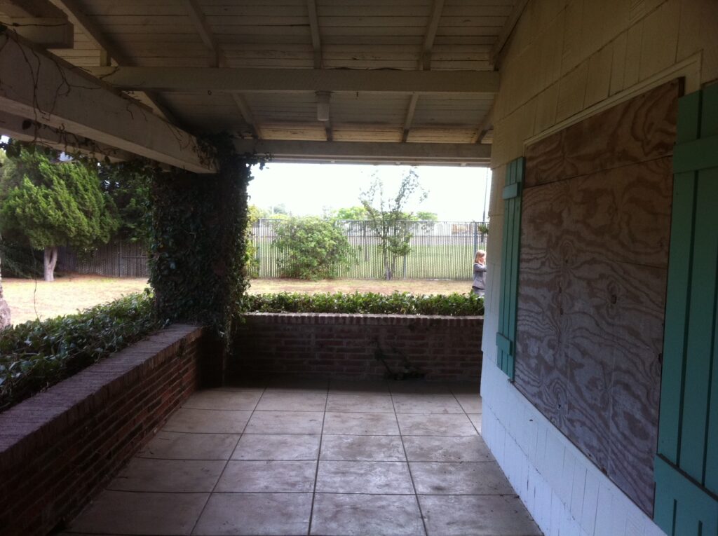 Covered porch with brick walls, green shutters, and tiled floor. View of a yard with trees, fence, and a person in the distance.
