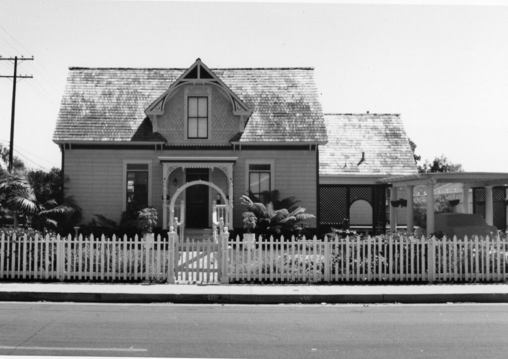 A black-and-white photograph of the Gerberding/Moranda Victorian house from the 1980s, featuring a shingled roof, white picket fence, and arched front porch with garden elements.
