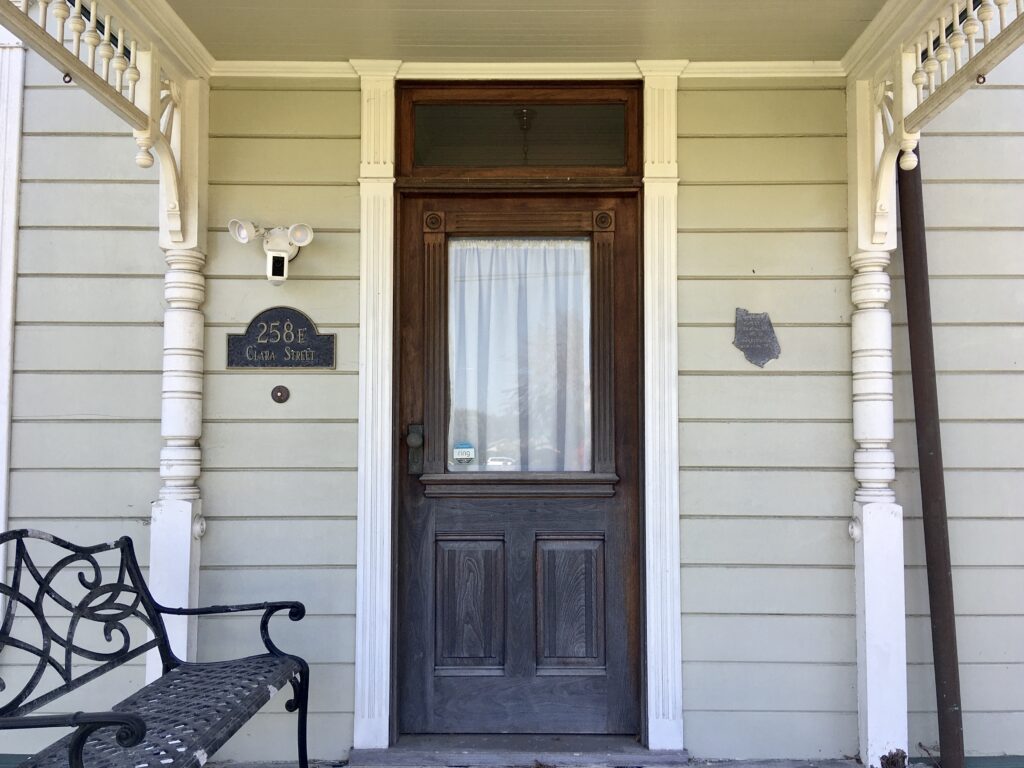 Front entrance of Gerberding/Moranda House at 258 E Clara Street, featuring a dark wooden door with transom window, white porch columns, and a black wrought iron bench on light beige siding.
