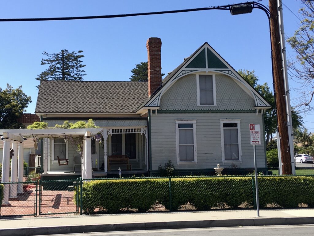 Light gray Victorian house with green trim, white porch columns, brick chimney, and "NO PARKING" sign, set against a blue sky with trees. Caption: Gerberding/Moranda House, 2022.