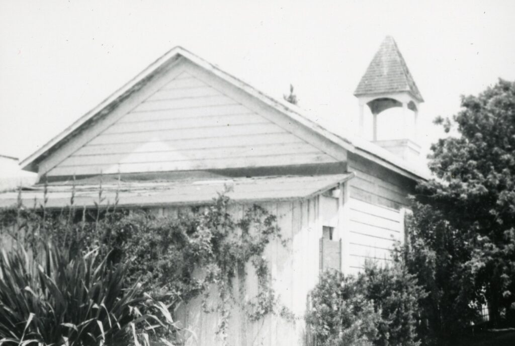 Black-and-white historical photograph of a wooden building with a gabled roof, small tower, and overgrown vegetation in the foreground.