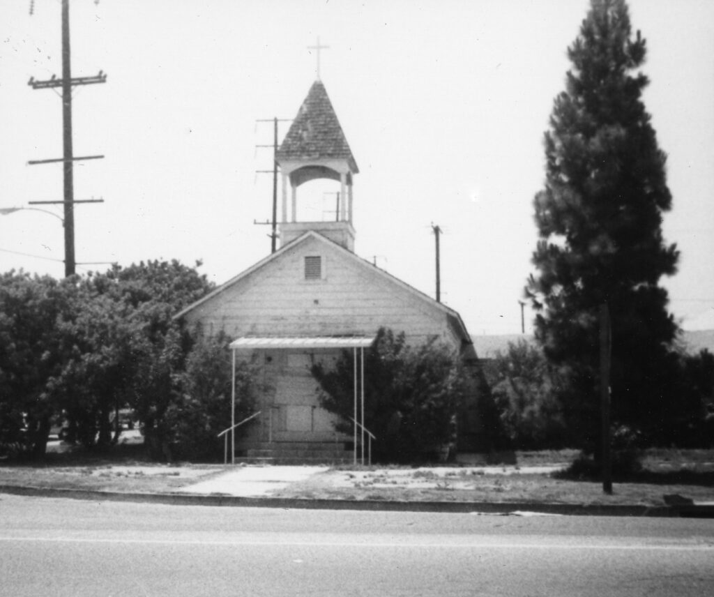 Black-and-white historical photograph of a small wooden church with a steeple and cross, situated along Wells Road, surrounded by trees and utility poles.