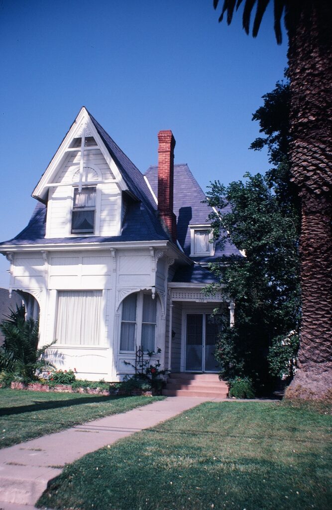 White Victorian house with dark blue roof and red brick chimney, surrounded by green lawn and palm trees. Photographed in 1982 as the Logan House.