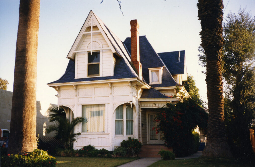 White Victorian house with dark blue shingles, dormer windows, and a brick chimney, surrounded by palm trees and landscaped gardens, photographed in 1985.
